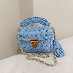 K&ouml;nnte beinhalten: Eine hellblaue geh&auml;kelte Handtasche mit einer goldenen Kettenkette und einem goldenen Verschluss. Die Tasche hat ein geflochtenes Design und einen Quastenanh&auml;nger.