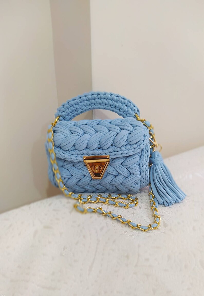 K&ouml;nnte beinhalten: Eine hellblaue geh&auml;kelte Handtasche mit einer goldenen Kettenkette und einem goldenen Verschluss. Die Tasche hat ein geflochtenes Design und einen Quastenanh&auml;nger.