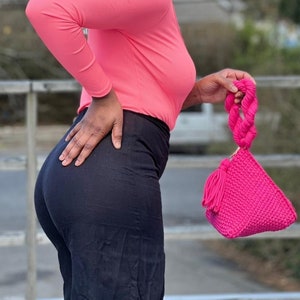 May include: A bright pink, triangular-shaped handbag with a braided handle and tassel detail. The bag is made of a textured knit material. The handle is a thick, twisted rope design. The bag is held by a person wearing a pink long-sleeve shirt and black pants.