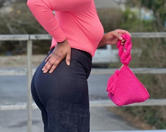 May include: A bright pink, triangular-shaped handbag with a braided handle and tassel detail. The bag is made of a textured knit material. The handle is a thick, twisted rope design. The bag is held by a person wearing a pink long-sleeve shirt and black pants.