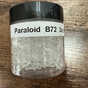 May include: A clear plastic jar with a black lid, filled with small, clear, irregular-shaped pieces. The label reads "Paraloid B72 3oz". The jar is sitting on a wooden surface.