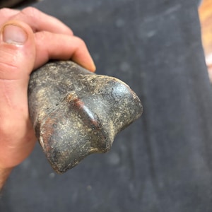 May include: A dark gray, weathered stone artifact with a rough, textured surface and a distinct, raised central ridge. The object has an irregular, elongated shape with earthy brown undertones visible along the edges and a dense, solid appearance.