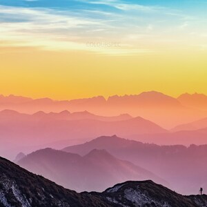 May include: A solitary figure stands on a mountain summit, silhouetted against a vibrant sunset. The sky transitions from blue to yellow, then orange, and finally fades into a soft pink over the distant mountain ranges.