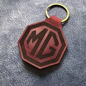 Op de afbeelding: Een rode leren sleutelhanger met een zwart gegraveerd MG-logo. De sleutelhanger heeft een gouden metalen ring.
