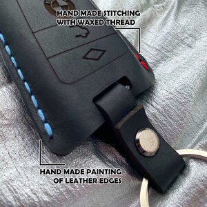 May include: Black leather key fob with blue stitching and a silver key ring. The fob has a hand-painted edge and the text "HAND MADE STITCHING WITH WAXED THREAD" is written on the top.