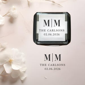 May include: A black and clear stamp with the initials "MM", the name "THE CARLSONS", and the date "02.06.2026" printed in black. The stamp is square and sits on a light pink and white background with floral accents.