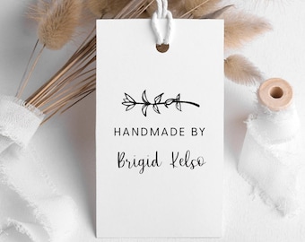 May include: A white tag with a black floral design and the text "HANDMADE BY Brigid Kelso". The tag is attached to a white string. Dried flowers and a spool of ribbon are in the background.