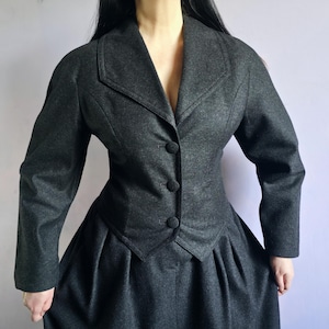 May include: A dark charcoal grey jacket and skirt set. The jacket features a notched lapel, long sleeves, and three fabric-covered buttons down the front. The skirt is high-waisted with deep pleats at the waist, creating a full, structured silhouette.