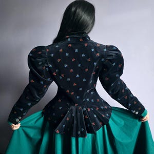 Puede incluir: Chaqueta de terciopelo azul marino oscuro con un estampado floral en rojo, azul y amarillo. La chaqueta tiene mangas abullonadas grandes y un detalle de peplum en la cintura. La chaqueta se combina con una falda verde turquesa.