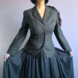 Pode incluir: Um blazer cinza escuro com colarinho de ponta e cinco botões. O blazer é ajustado e tem a cintura cintada. O blazer é usado sobre uma saia de bolinhas pretas e brancas.