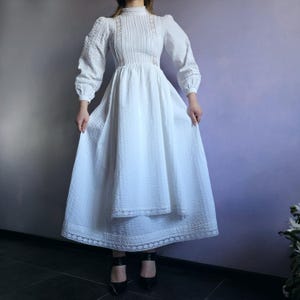 May include: A white long-sleeved dress featuring a high neckline, vertical pleating on the bodice, and intricate lace trim detailing along the sleeves and hem. The garment has a fitted waist and a full, flowing skirt with a textured fabric finish.