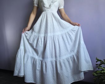 Puede incluir: Un vestido blanco de ojal con mangas cortas y una falda escalonada. El vestido presenta un escote cuadrado con un cuello con volantes y un corpiño ajustado. El vestido es largo hasta el suelo y tiene una silueta femenina.