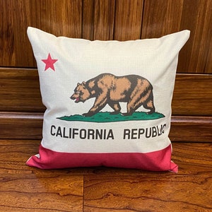 May include: A white cushion with a red, white and green California state flag design. The flag features a brown bear, a red star and the words "CALIFORNIA REPUBLIC".