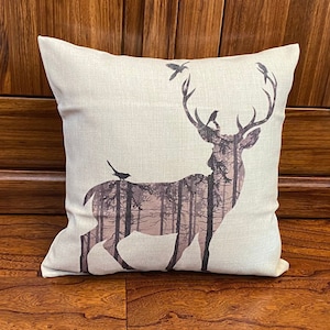 Könnte beinhalten: Ein beiger Kissenbezug mit einem bedruckten Design einer Hirschsilhouette mit einer Waldszene im Inneren. Der Hirsch hat Geweih und kleine Vögel sitzen auf dem Geweih und dem Rücken des Hirsches.