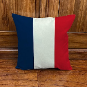 Pode incluir: Almofada quadrada com o design da bandeira francesa. A almofada tem listras verticais em azul, branco e vermelho. A almofada é feita de um tecido texturizado e é exibida em um fundo de madeira.