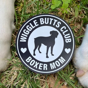 May include: A black and white 3-inch round magnet with the text "Wiggle Butts Club Boxer Mom" and a silhouette of a Boxer dog. The magnet is on a bed of green grass.