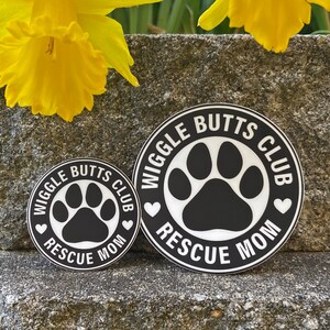 May include: Two black and white round stickers with the text "Wiggle Butts Club Rescue Mom" and a paw print in the center. The larger sticker is on top of the smaller sticker.