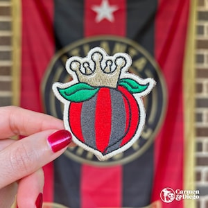 May include: A red and black embroidered patch of a peach with a gold crown on top. The peach has a green leaf on each side. The patch is in front of a red and black striped background.