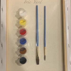 May include: A paint by numbers kit with the title "Irie Yard" on a canvas. The kit includes six small paint pots with white, yellow, red, blue, brown, and black paint. Two paintbrushes are included, one with a flat brush head and one with a round brush head.