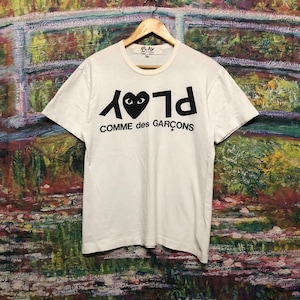 Puede incluir: Camiseta blanca con un logotipo negro de Play Comme des Gar&ccedil;ons. El logotipo presenta un coraz&oacute;n negro con ojos y la palabra "Play" escrita al rev&eacute;s.