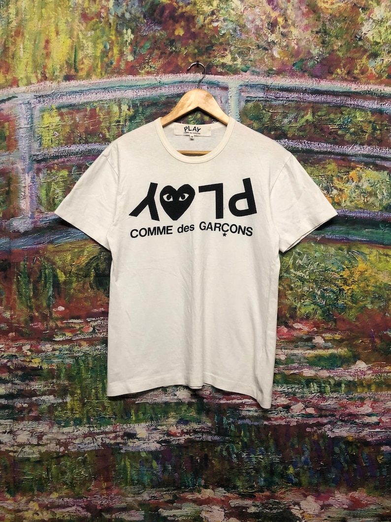 Puede incluir: Camiseta blanca con un logotipo negro de Play Comme des Gar&ccedil;ons. El logotipo presenta un coraz&oacute;n negro con ojos y la palabra "Play" escrita al rev&eacute;s.