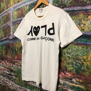 Puede incluir: Camiseta blanca con un logotipo negro de Play Comme des Gar&ccedil;ons que presenta un coraz&oacute;n con ojos. El logotipo est&aacute; invertido y el texto est&aacute; en may&uacute;sculas.