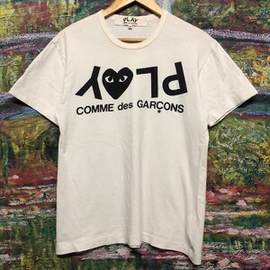 Puede incluir: Camiseta blanca con un logotipo negro de Play Comme des Gar&ccedil;ons. El logotipo presenta un coraz&oacute;n con ojos y el texto "Play" escrito al rev&eacute;s.