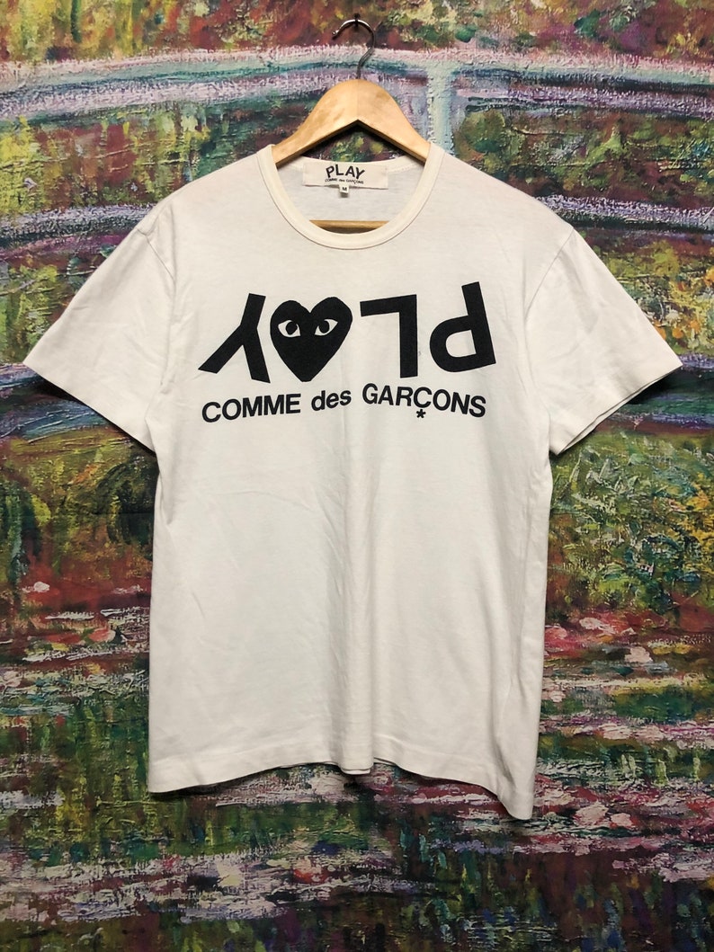 Puede incluir: Camiseta blanca con un logotipo negro de Play Comme des Gar&ccedil;ons. El logotipo presenta un coraz&oacute;n con ojos y el texto "Play" escrito al rev&eacute;s.