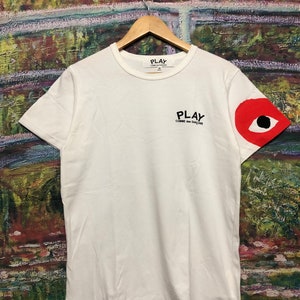 Peut inclure: T-shirt blanc avec un motif d'&oelig;il rouge sur la manche droite. Le t-shirt porte l'inscription "PLAY COMME des GARCONS" imprim&eacute;e sur le devant.