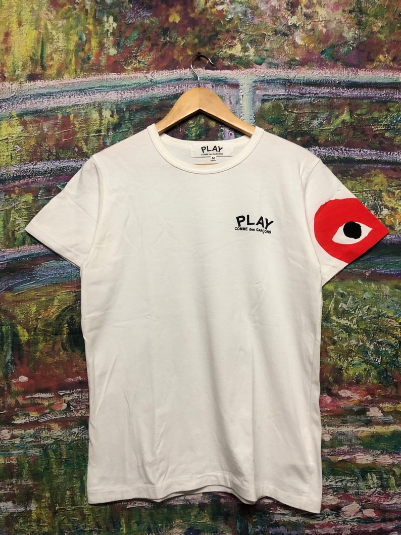 Peut inclure: T-shirt blanc avec un motif d'&oelig;il rouge sur la manche droite. Le t-shirt porte l'inscription "PLAY COMME des GARCONS" imprim&eacute;e sur le devant.