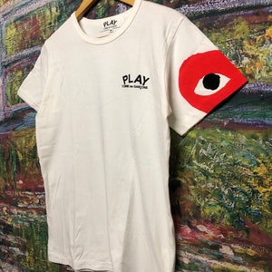 Peut inclure: T-shirt blanc avec un logo noir "PLAY COMME des GARCONS" sur le devant et un motif d'&oelig;il rouge sur la manche droite.