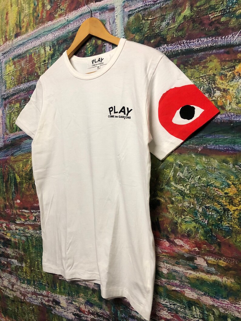 Peut inclure: T-shirt blanc avec un logo noir "PLAY COMME des GARCONS" sur le devant et un motif d'&oelig;il rouge sur la manche droite.