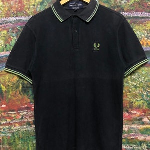 Puede incluir: Polo negro de manga corta con cuello y puños a rayas verdes y blancas. La camisa presenta un pequeño logotipo bordado de una corona de laurel con las letras "CDG" en su interior.