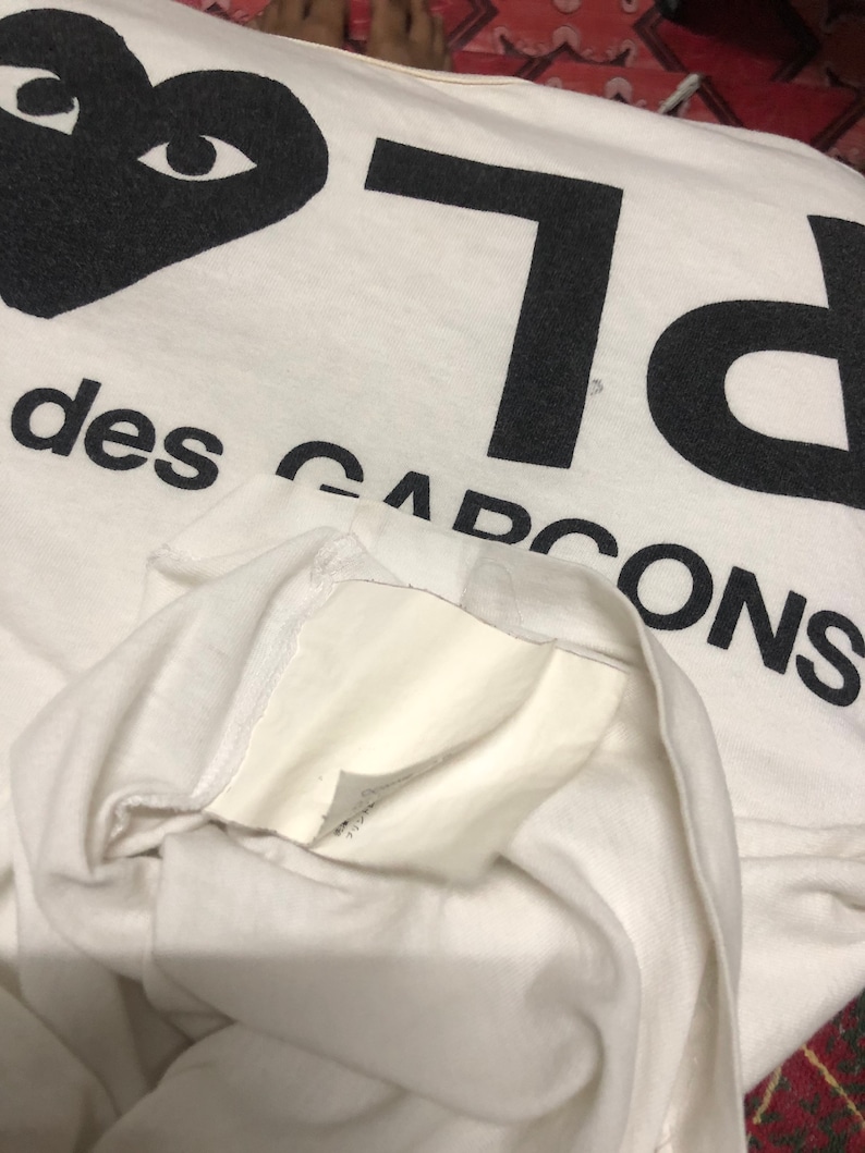 Puede incluir: Camiseta blanca con un coraz&oacute;n negro con ojos y el texto "PLAY" y "des GARCONS" impreso en ella.