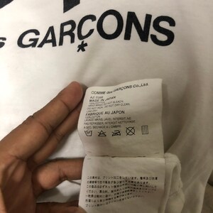 Puede incluir: Camiseta blanca con texto negro que dice "COMME des GARCONS*" y una etiqueta de cuidado en blanco y negro con instrucciones para lavar y secar la camiseta. El texto de la etiqueta de cuidado dice "COMME des GARCONS Co., Ltd. AZ-T068 MADE IN JAPAN 100% COTTON HAND WASH DO NOT BLEACH DO NOT IRON DO NOT DRY CLEAN. LINE DRY FABRIQUE AU JAPON 100% COTON LAVAGE MAINS. JAVEL INTERDIT. NE PAS REPASSER, INTERDIT NETTOYAGE A SEC. SECHAGE A L'OMBRE."