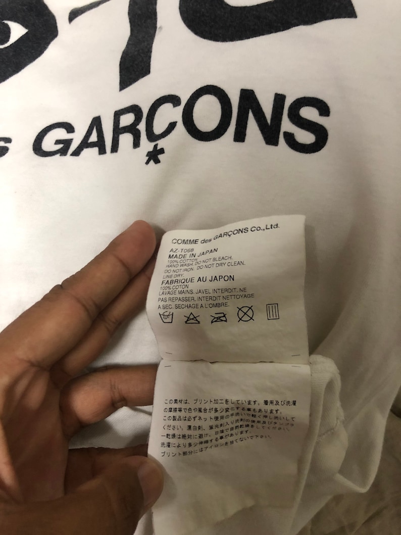Puede incluir: Camiseta blanca con texto negro que dice "COMME des GARCONS*" y una etiqueta de cuidado en blanco y negro con instrucciones para lavar y secar la camiseta. El texto de la etiqueta de cuidado dice "COMME des GARCONS Co., Ltd. AZ-T068 MADE IN JAPAN 100% COTTON HAND WASH DO NOT BLEACH DO NOT IRON DO NOT DRY CLEAN. LINE DRY FABRIQUE AU JAPON 100% COTON LAVAGE MAINS. JAVEL INTERDIT. NE PAS REPASSER, INTERDIT NETTOYAGE A SEC. SECHAGE A L'OMBRE."