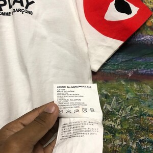 Peut inclure: T-shirt blanc avec un c&oelig;ur rouge avec un &oelig;il noir sur la manche droite. Le t-shirt porte l'inscription "PLAY COMME des GAR&Ccedil;ONS" sur le devant. L'&eacute;tiquette d'entretien est visible et comprend des instructions pour le lavage et le s&eacute;chage du t-shirt. Le texte sur l'&eacute;tiquette est le suivant : "COMME des GAR&Ccedil;ONS Co., Ltd. AZ-T095 MADE IN JAPAN 100% COTTON HAND WASH, DO NOT BLEACH. DO NOT IRON, DO NOT DRY CLEAN. LINE DRY, FABRIQUE AU JAPON 100% COTON LAVAGE MAINS. JAVEL INTERDIT NE PAS REPASSER INTERDIT NETTOYAGE SEC. SECHAGE AL'OMBRE."