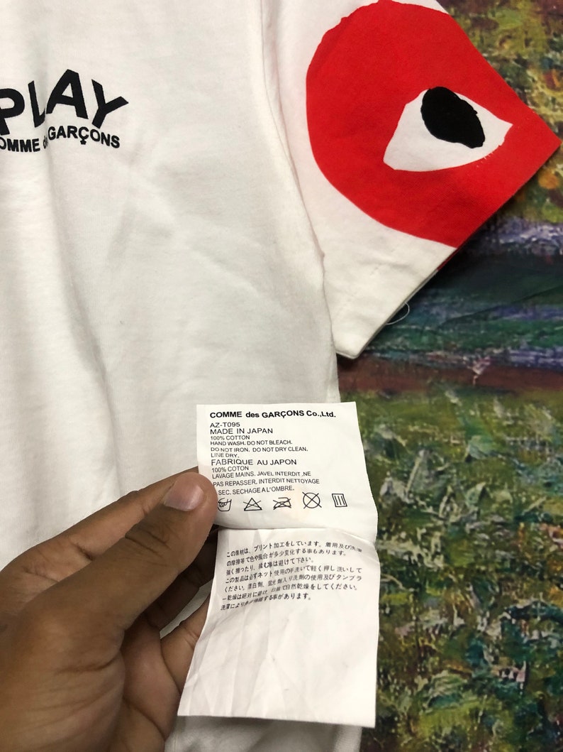 Peut inclure: T-shirt blanc avec un c&oelig;ur rouge avec un &oelig;il noir sur la manche droite. Le t-shirt porte l'inscription "PLAY COMME des GAR&Ccedil;ONS" sur le devant. L'&eacute;tiquette d'entretien est visible et comprend des instructions pour le lavage et le s&eacute;chage du t-shirt. Le texte sur l'&eacute;tiquette est le suivant : "COMME des GAR&Ccedil;ONS Co., Ltd. AZ-T095 MADE IN JAPAN 100% COTTON HAND WASH, DO NOT BLEACH. DO NOT IRON, DO NOT DRY CLEAN. LINE DRY, FABRIQUE AU JAPON 100% COTON LAVAGE MAINS. JAVEL INTERDIT NE PAS REPASSER INTERDIT NETTOYAGE SEC. SECHAGE AL'OMBRE."
