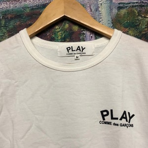 Peut inclure: T-shirt blanc avec un logo noir PLAY COMME des GAR&Ccedil;ONS sur le devant. Le t-shirt a un col rond et des manches courtes. L'&eacute;tiquette sur le t-shirt indique PLAY COMME des GAR&Ccedil;ONS et la taille M.