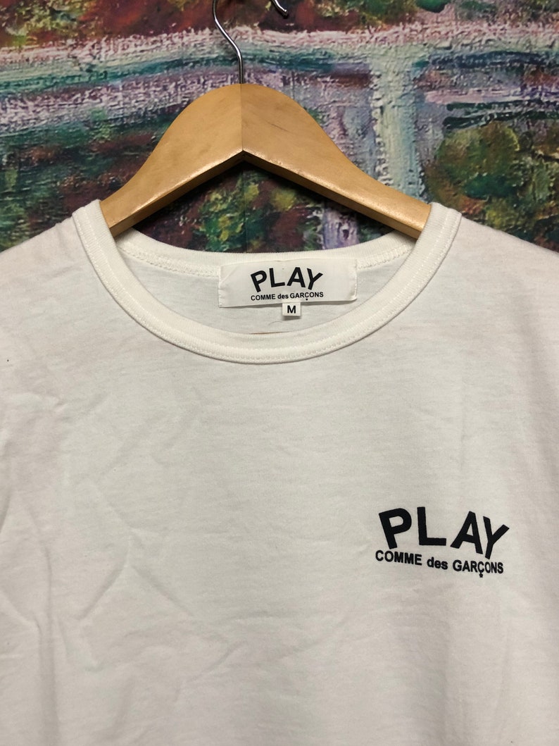 Peut inclure: T-shirt blanc avec un logo noir PLAY COMME des GAR&Ccedil;ONS sur le devant. Le t-shirt a un col rond et des manches courtes. L'&eacute;tiquette sur le t-shirt indique PLAY COMME des GAR&Ccedil;ONS et la taille M.