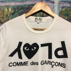 Puede incluir: Camiseta blanca con un logotipo negro de Play Comme des Gar&ccedil;ons que presenta un coraz&oacute;n con ojos y el texto "Play" y "Comme des Gar&ccedil;ons" en una fuente estilizada.