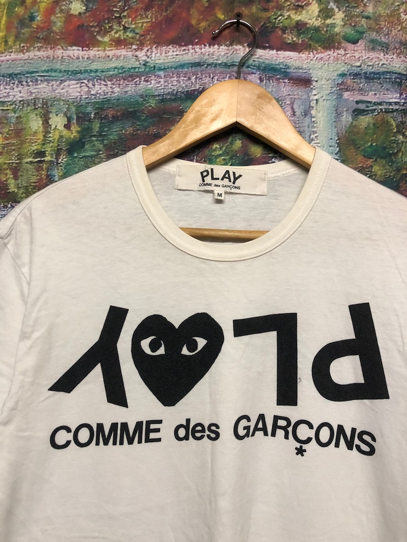 Puede incluir: Camiseta blanca con un logotipo negro de Play Comme des Gar&ccedil;ons que presenta un coraz&oacute;n con ojos y el texto "Play" y "Comme des Gar&ccedil;ons" en una fuente estilizada.