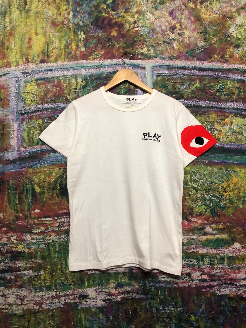 Peut inclure: T-shirt blanc avec un motif d'&oelig;il rouge sur la manche gauche. Le t-shirt porte l'inscription "PLAY" en noir sur le devant, avec les mots "COMME des GARCONS" en noir plus petit en dessous.
