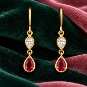 May include: A pair of gold drop earrings featuring a small pear-shaped clear stone above a larger pear-shaped red stone. Each earring hangs from a simple gold hook, showcasing a polished metal setting that secures both faceted gemstones.