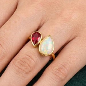 May include: A gold ring featuring two pear-shaped stones set in a bypass style. One stone is a deep red ruby, and the other is a white iridescent opal. The band is thin and polished, showcasing a minimalist design that highlights the contrasting colors.