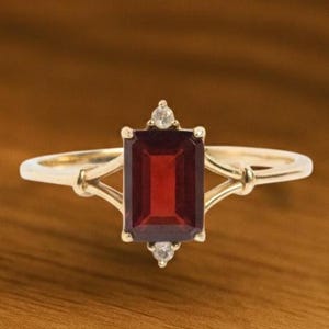 May include: A gold ring featuring a rectangular, deep red gemstone. The ring has two smaller, clear gemstones above and below the main stone. The band has an intricate design.