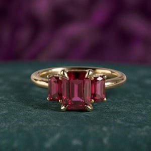Op de afbeelding: Een gouden ring met drie rechthoekige, dieprode edelstenen. De centrale steen is groter dan de twee aan weerszijden. De ring ligt op een donkergroen oppervlak met een vage paarse achtergrond.