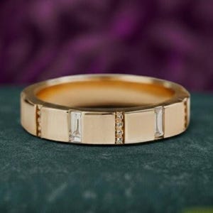 May include: A gold band ring with rectangular and small diamond accents. The ring has a flat design with a polished finish. The ring is set against a dark green surface with a blurred purple background.
