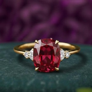 Könnte beinhalten: Ein Goldring mit einem großen, tiefroten Edelstein im Kissenschliff in der Mitte, flankiert von zwei kleineren, klaren Steinen im Rundschliff auf jeder Seite. Der Ring besteht aus poliertem Gelbgold und hält die Steine in einer Krappenfassung.