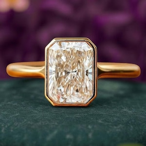 Puede incluir: Un anillo de oro con una gema grande, rectangular y transparente. La gema está engastada en un bisel de oro y tiene un corte brillante. La banda del anillo también es de oro y tiene un acabado liso y pulido. El fondo es borroso, morado y verde.